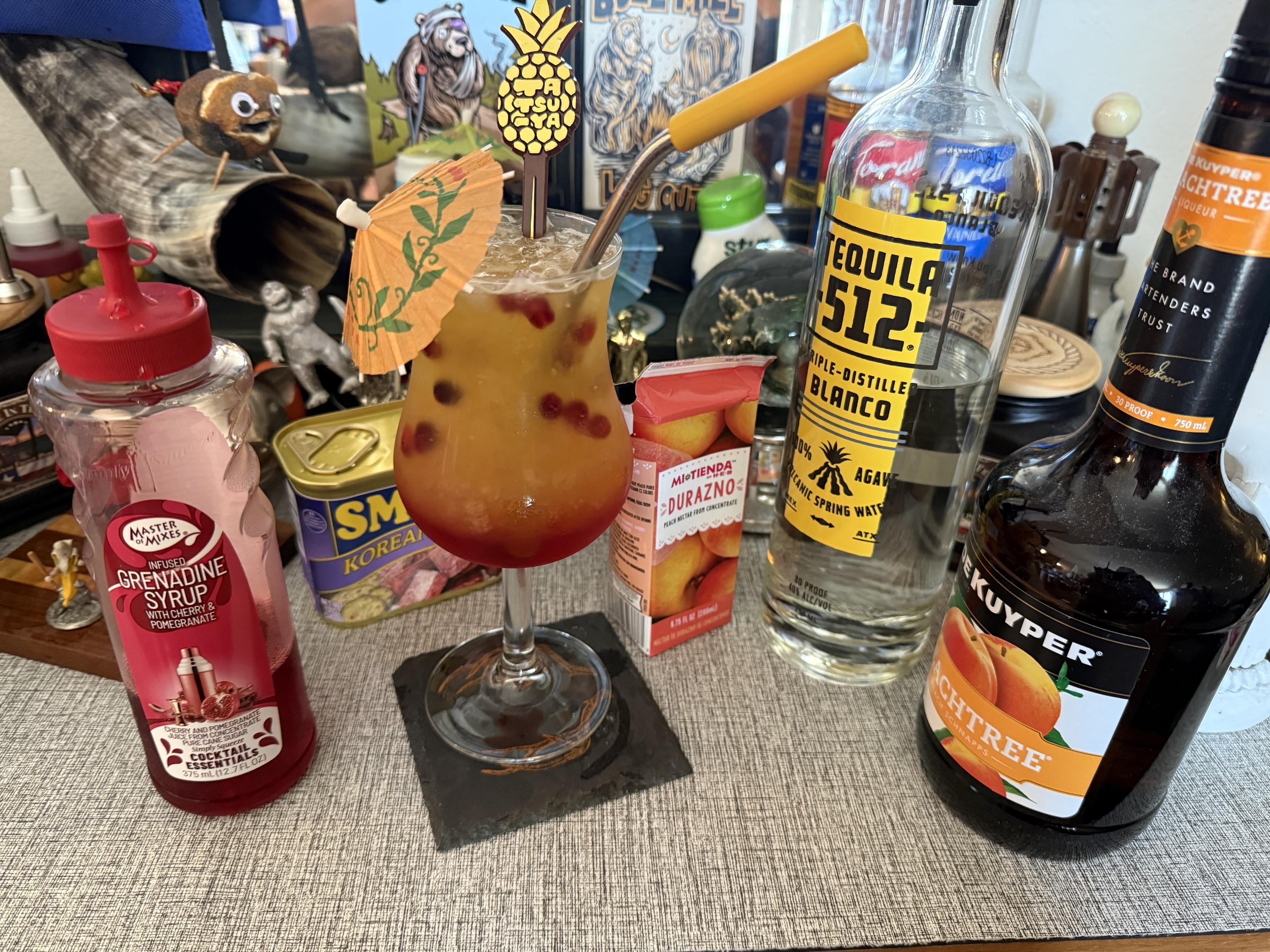 A colorful cocktail sits in a glass with an umbrella and garnishes. Nearby are bottles of tequila, peach schnapps, and grenadine syrup, along with a carton of peach nectar. The background features various decorative items and figurines. Tequila Sunrise  variant, recipe in parent toot!